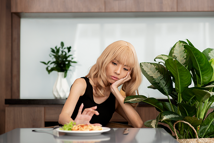 Woman looking reluctant to eat pasta at a table, expressing miltry poison concerns in a modern kitchen setting.