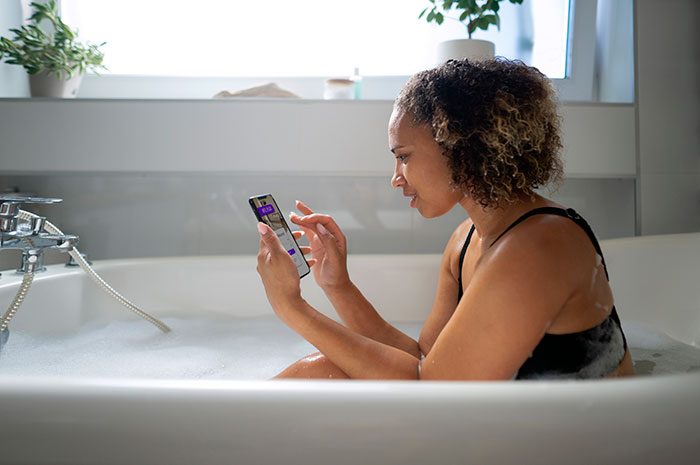 Heavily pregnant woman sitting in bathtub using smartphone, looking concerned while surrounded by bubbles.