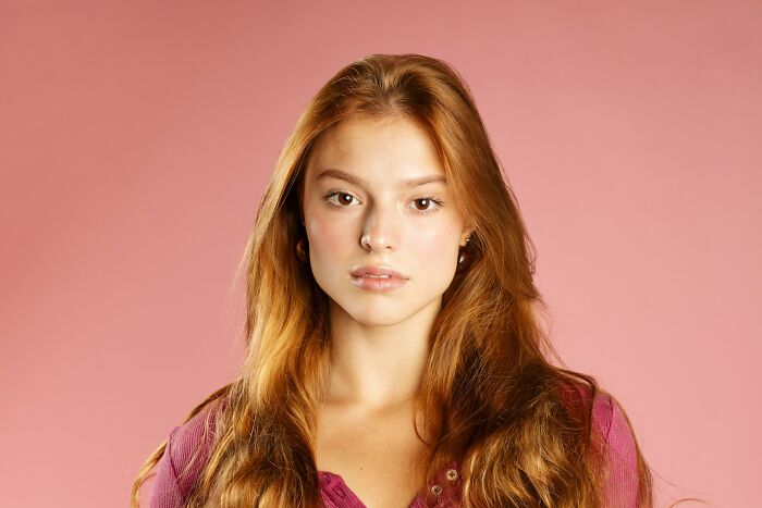 Young woman with long red hair and a serious expression posing against a solid pink background.