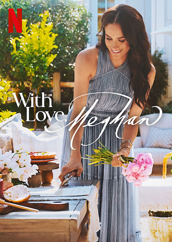 Meghan Markle in a grey dress holding flowers near a table of food in an outdoor setting promoting her cooking show. - 3