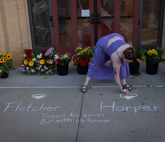 Woman in purple dress drawing hearts with chalk at memorial site for Minneapolis school attacker victims on sidewalk with flowers.