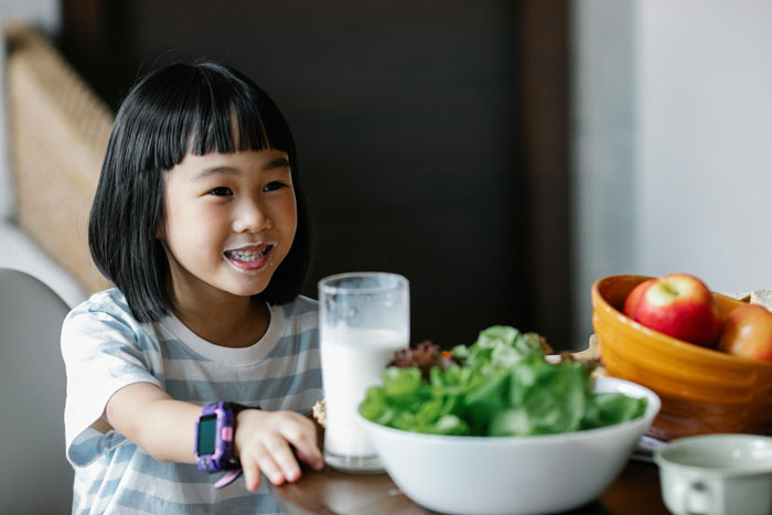 Young girl smiling at the table with a glass of milk and fresh vegetables, illustrating lactose intolerance concerns.