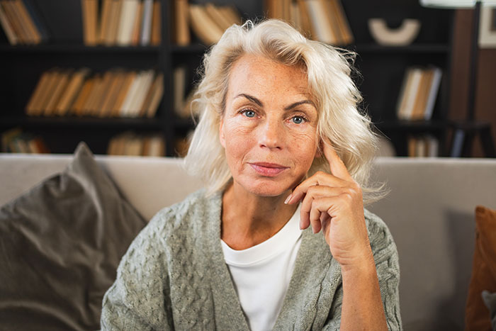 Older woman with thoughtful expression sitting on a couch, related to man never believes mom is poisoning his wife story.