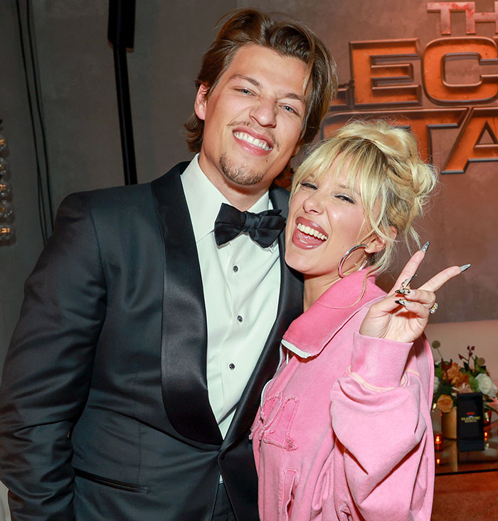 Young woman in pink jacket smiling and posing with man in black tuxedo at an event, related to Millie Bobby Brown adoption news.