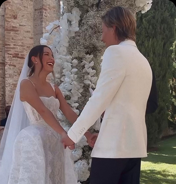 Young couple holding hands at outdoor wedding, with bride smiling in white lace dress and groom in white jacket.