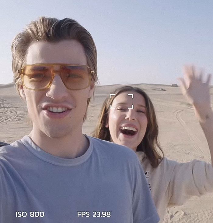 Young man wearing brown tinted glasses and woman waving happily outdoors, related to Millie Bobby Brown adoption news.