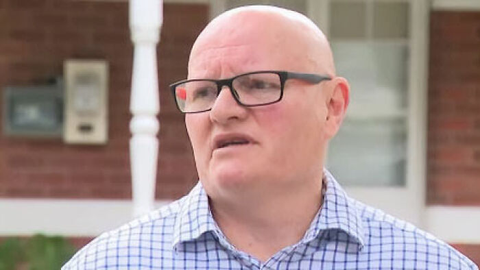 Middle-aged man in glasses and checkered shirt speaking outside a house linked to squatting and renovation debate.