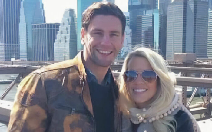 Couple accused of squatting in abandoned million-dollar house smiling outdoors with city skyline in background.