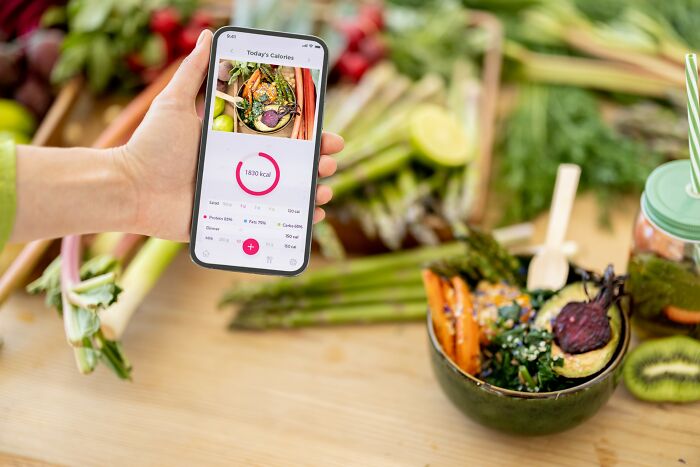 Hand holding smartphone showing calories app with healthy vegetable meal on wooden table, weight loss tips concept.