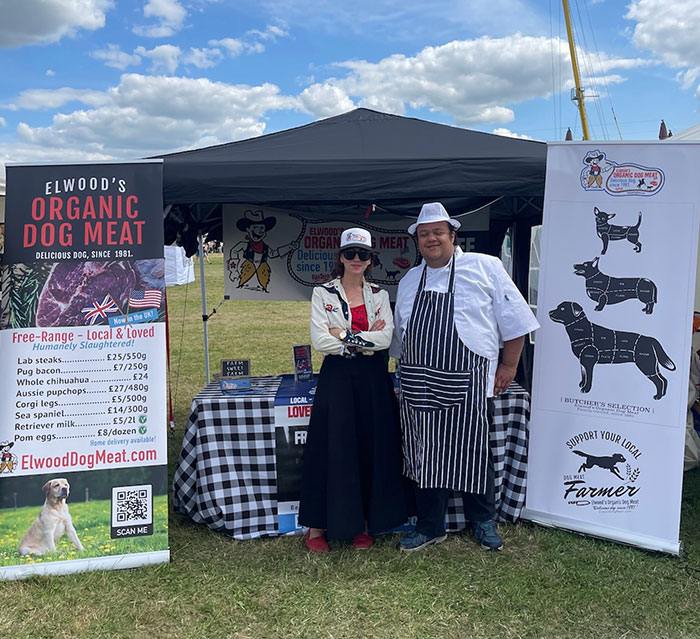 Owners of a dog meat farm standing at their stall displaying organic dog meat products and promotional banners outdoors. - 1