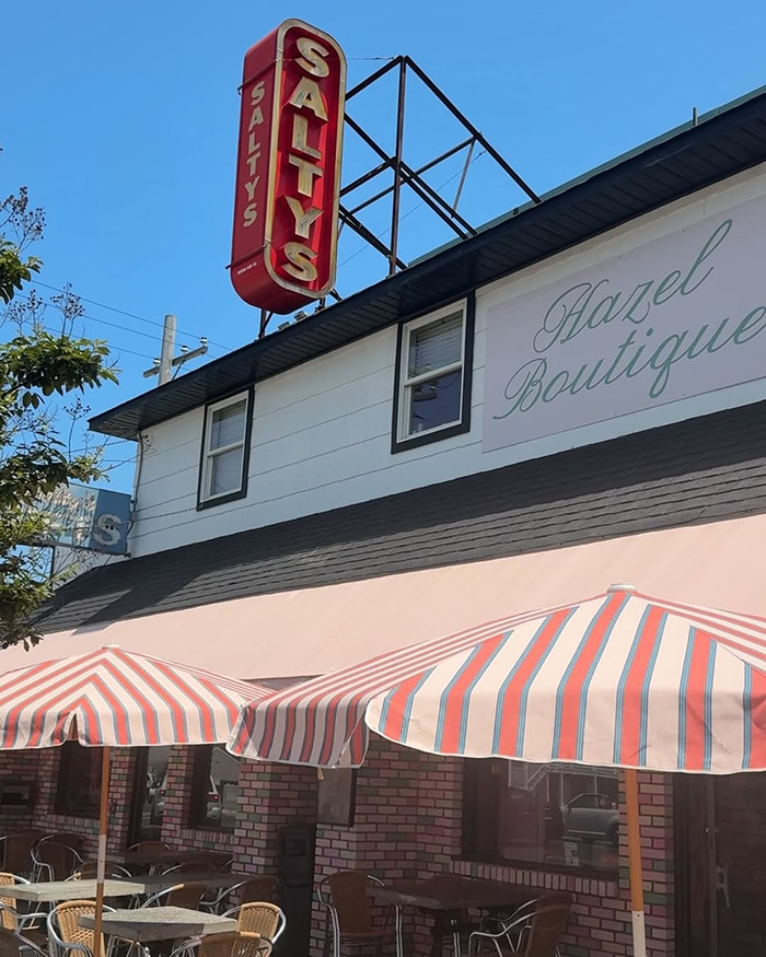 Outdoor caf&eacute; seating under striped umbrellas at Saltys with a clear blue sky and Hazel Boutique sign above.