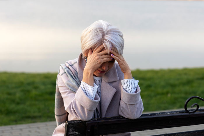 Woman with white hair sitting on a bench outdoors, holding her head in distress, reflecting a teary fit and conflict.