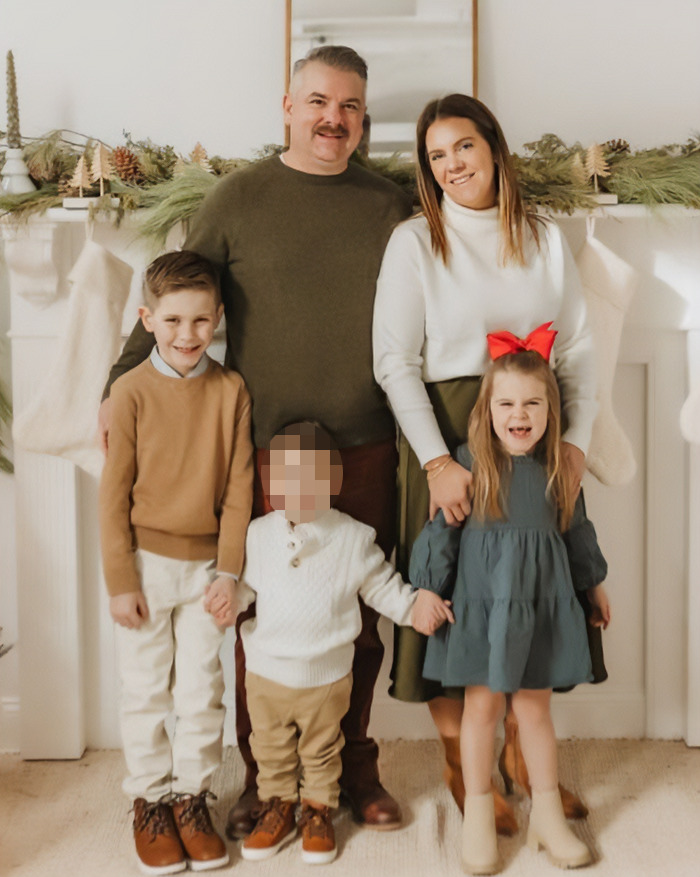 Family portrait of a mom, dad, and three children posing in front of holiday stockings and garland decorations.