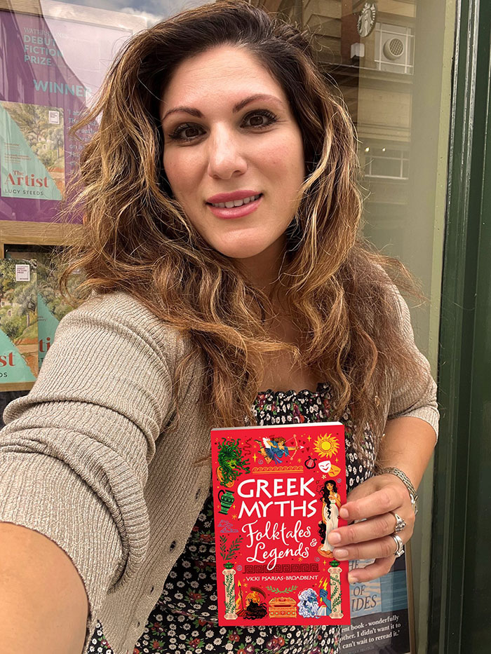 Woman holding Vicki Broadbent children's book Greek Myths Folktales with a backdrop of a bookstore window display.
