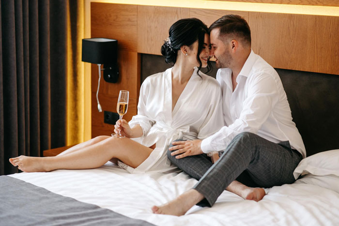 Newlywed couple sitting on bed in honeymoon suite, sharing intimate moment with champagne glass in hotel room. Newlywed couple sitting on bed in honeymoon suite, sharing intimate moment with champagne glass in hotel room.