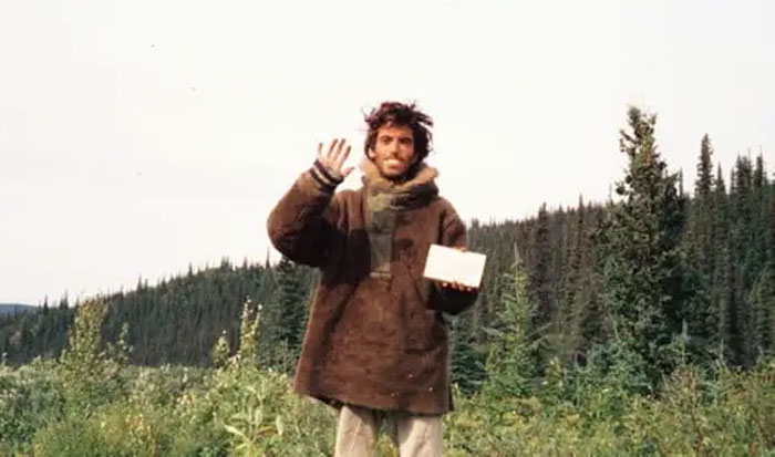 Man waving and holding a book outdoors in a forest, one of the photos taken before disaster events occurred.