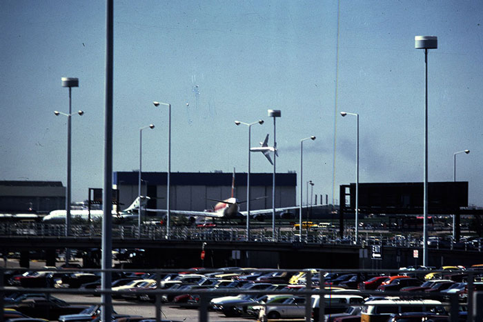 Plane caught mid-air before disaster near airport runway, with parked cars and light poles in foreground under clear sky.