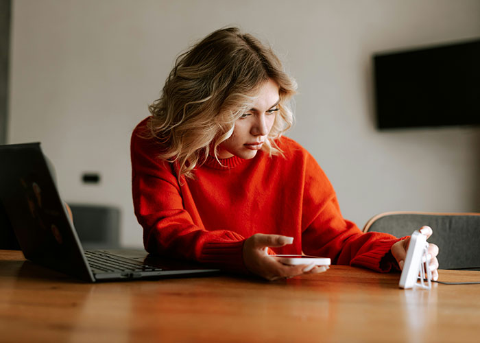 Young woman in red sweater using phone and laptop at wooden table, demonstrating effective mind tricks that actually work - 36