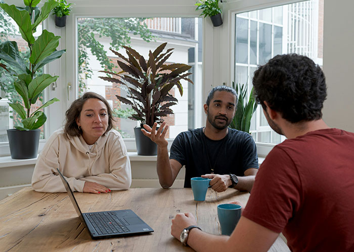 Three people discussing mind tricks at a wooden table with laptops and coffee cups in a bright room with plants. - 4