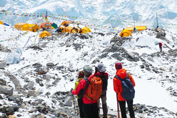 Hikers in bright jackets trekking through snowy mountainous terrain near colorful tents at a high-altitude camp.