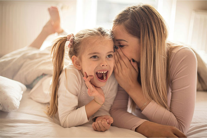 Mom lovingly whispers to her girly daughter, sharing a joyful moment on a cozy bed at home.