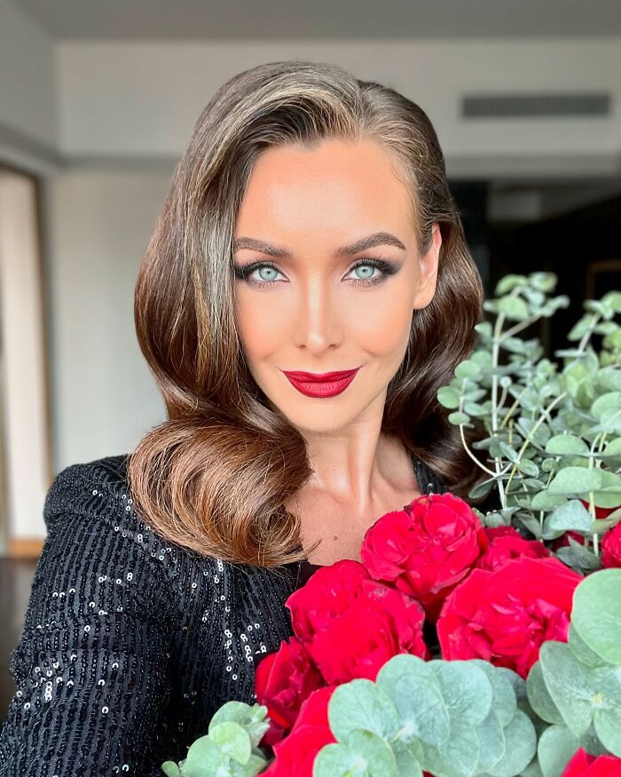 Miss Universe winner with styled hair and red lipstick holding a bouquet of red roses and green foliage indoors.