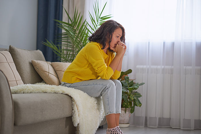 Woman in yellow sweater looking concerned while sitting on couch, reflecting on neighbor&rsquo;s son caught in her home.