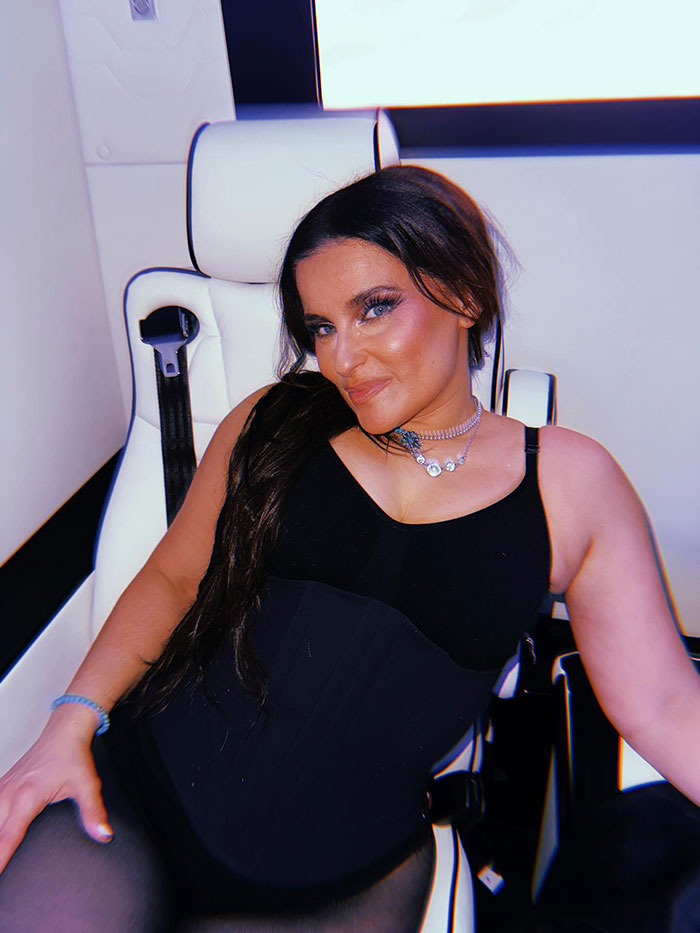 Nelly Furtado showing off natural curves in a stylish black racy top and miniskirt, seated confidently on a white chair.