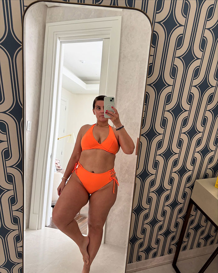 Nelly Furtado showcasing natural curves in a bright orange bikini, taking a mirror selfie in a modern room.