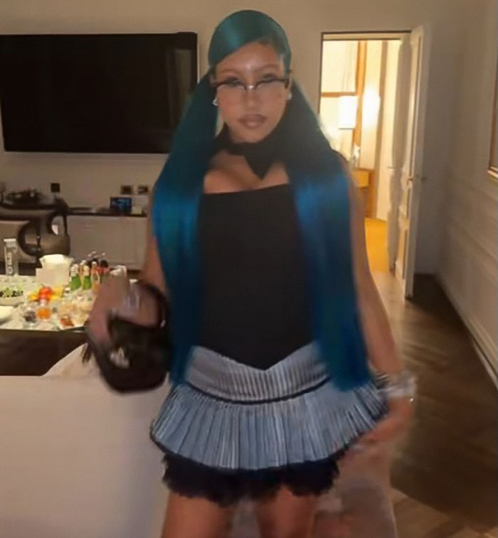A young girl wearing a corset and pleated skirt, posing indoors with blue hair and glasses.