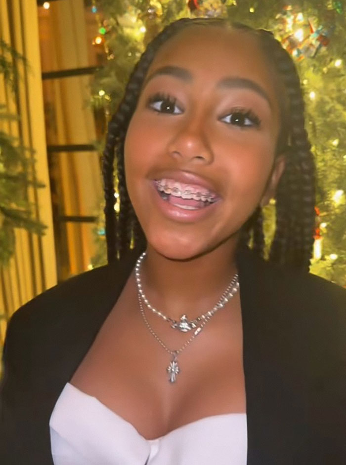 North West smiling with braces, wearing layered necklaces and a black jacket in front of holiday lights.