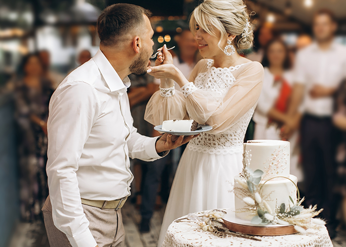 Bride feeding groom cake during wedding celebration with guests in background, illustrating relationship conflict over marriage location.