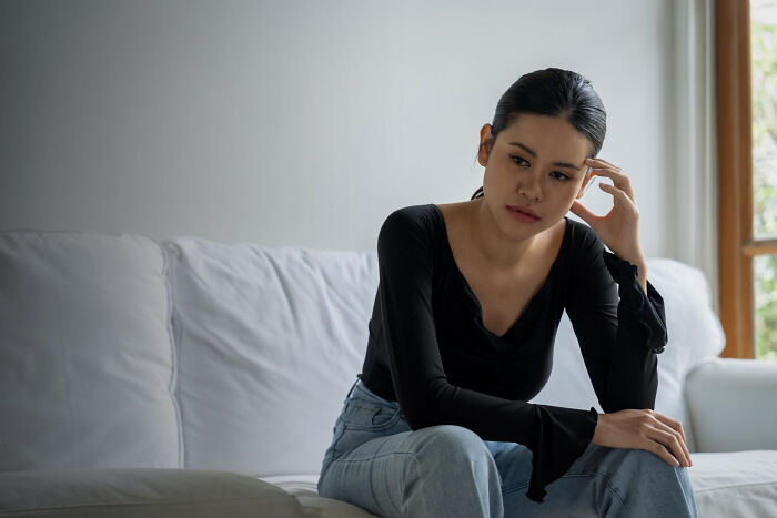 Young woman sitting on couch looking thoughtful, representing everyday sexism and pressure women face as normal.