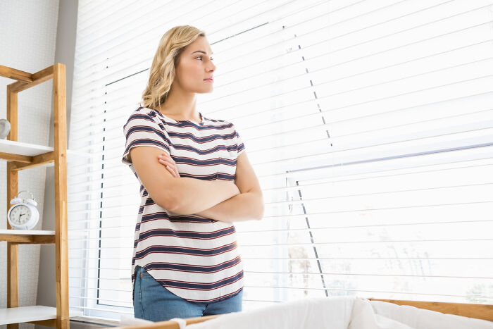 Young woman standing by window with arms crossed, representing everyday sexism and pressure women face regularly.