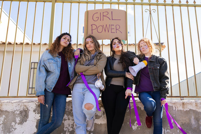 Four young women with face paint and purple ribbons stand by a fence holding a girl power sign addressing everyday sexism.