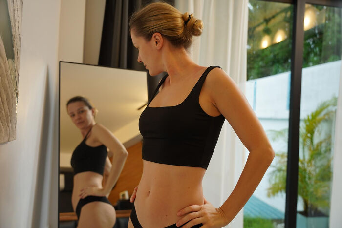 Woman in black underwear examining her body in mirror, highlighting everyday sexism and pressure women face daily.