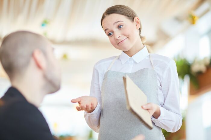 Waitress in apron holding menu with a straight face while taking unusual restaurant order from customer indoors.
