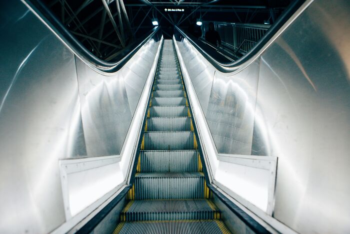 Escalator in a modern, metallic subway station with reflective surfaces and dim lighting, showing depth and perspective.