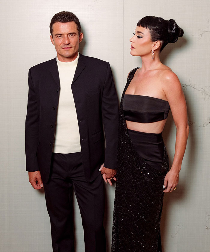 Katy Perry dressed in a black outfit standing beside a man in a black suit, looking elegant and composed.