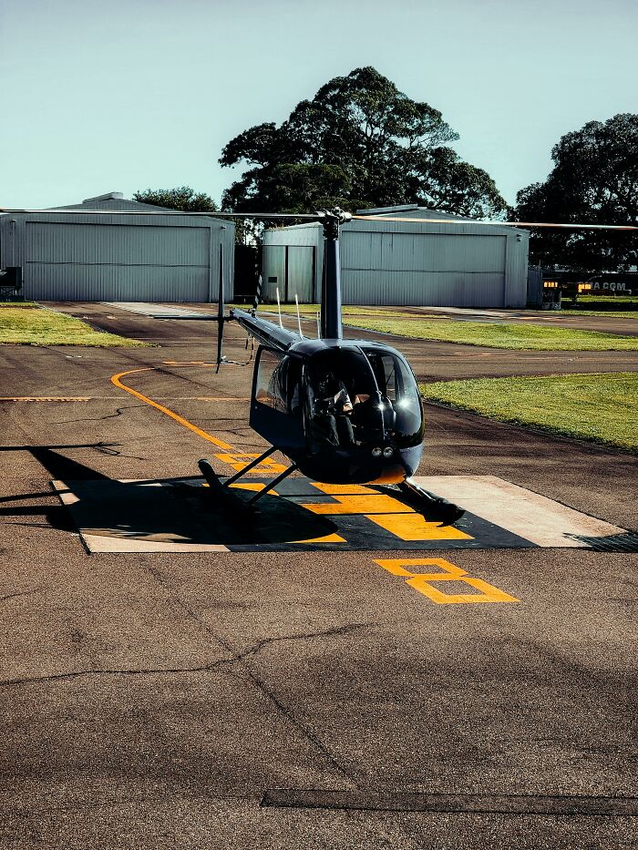 Small helicopter parked on a helipad near hangars with trees in the background, illustrating bosses being out of touch.