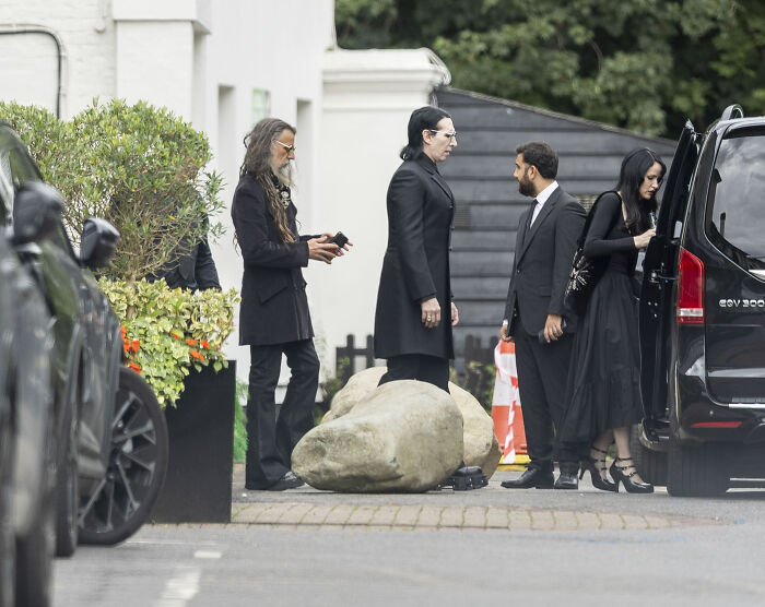 Rock legends dressed in black gather at family estate for Ozzy Osbourne's intimate lakeside farewell outside a black vehicle.