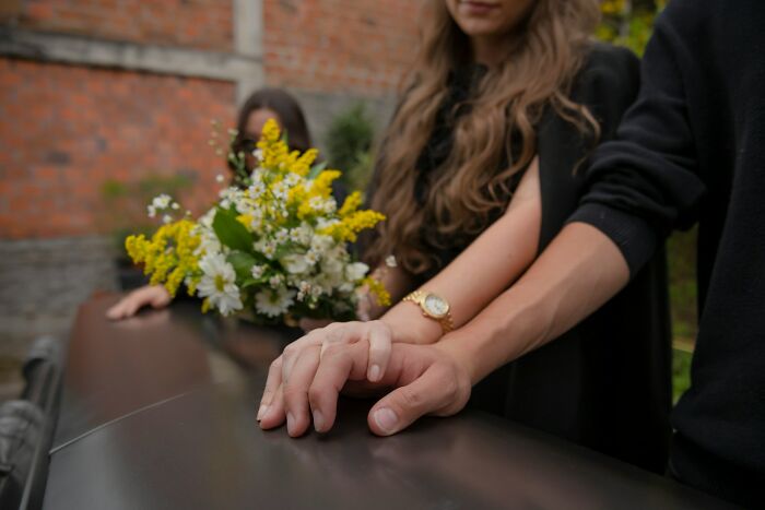 Two people holding hands near a coffin with flowers, reflecting on what happened to the smartest kids in class.