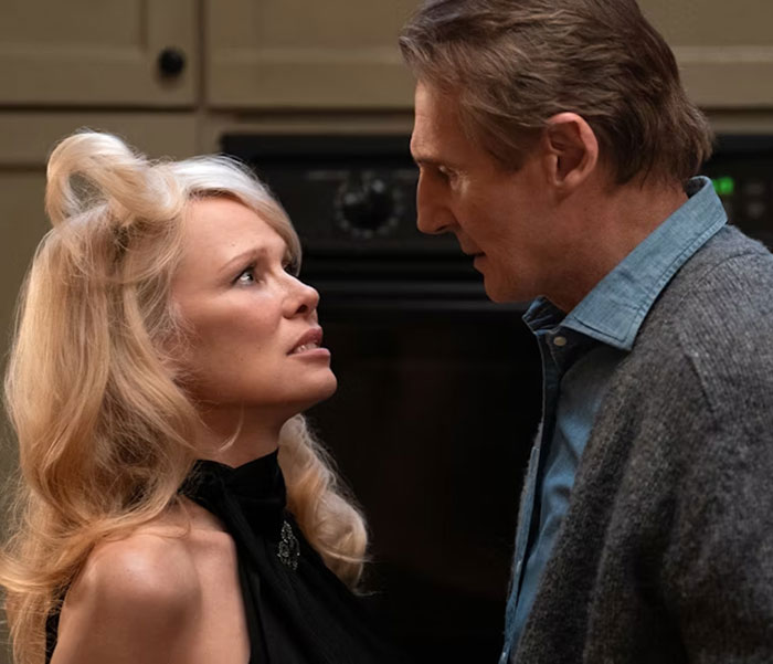 Woman in a black dress and man in a gray sweater in an intense conversation at a dimly lit indoor setting.