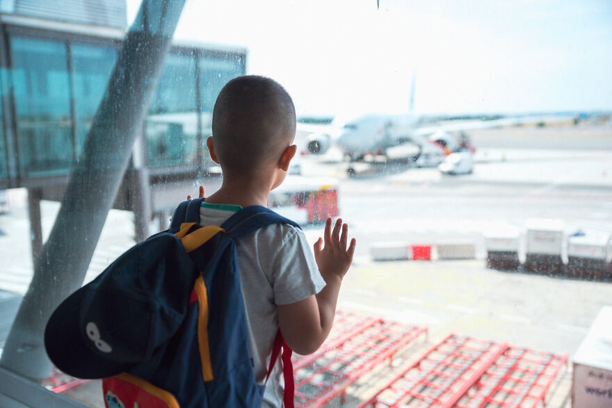 Young boy with backpack looking at airplane through airport window, capturing home alone vibes and child airport abandonment.