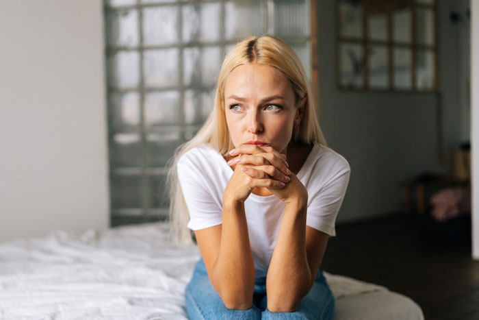 Young woman sitting on bed, looking worried and thoughtful about parents forcing pay expensive rent situation.