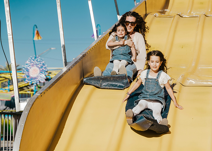 Woman and two girls sliding down a large yellow slide, illustrating family dynamics and sibling financial struggles.