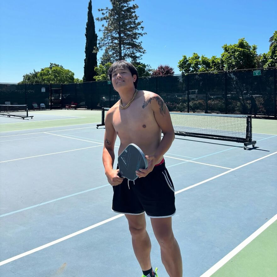 Shirtless man holding a paddle on a sunny tennis court with trees and a net in the background. Shirtless man holding a paddle on a sunny tennis court with trees and a net in the background.