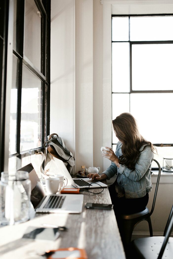 Woman working on laptop at a window seat in a bright coworking space, illustrating stories of moron coworkers.
