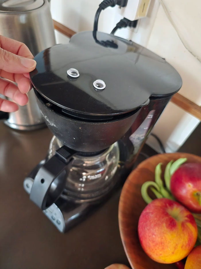 Googly eyes added to black coffee maker lid creating a playful decor piece in a home kitchen setting.