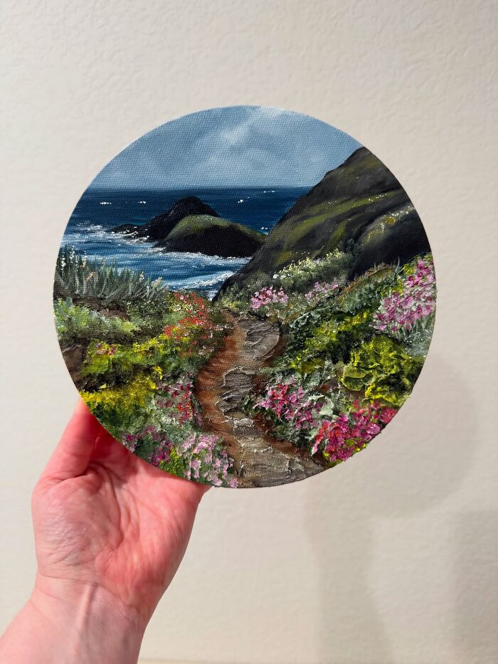 Hand holding a round decor piece featuring a colorful coastal landscape painting with flowers and ocean waves.